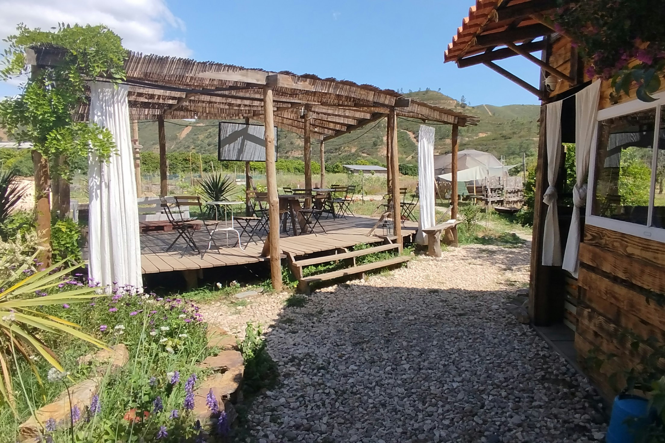 Campingplatz mit Kiesweg, Holzterrasse und Pergola, Blick auf Zäune und Berge im Hintergrund.