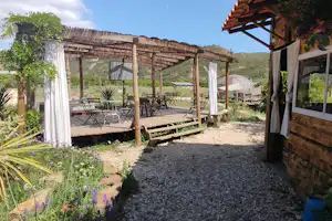 Campingplatz mit Kiesweg, Holzterrasse und Pergola, Blick auf Zäune und Berge im Hintergrund.