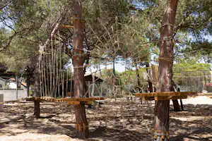 Holzpfosten und Seile an einem Baum, daneben ein Campingplatzweg mit Sitzgelegenheiten und Zäunen im Grünen.