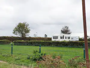Wohnmobil hinter einem grünen Zaun, daneben Hecken und Bäume, im Hintergrund ein Feldweg.