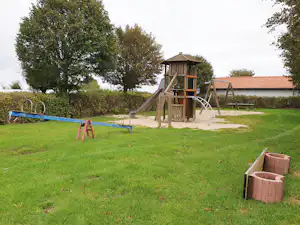 Spielplatz am Campingplatz mit Holz-Spielturm, Rutsche, Schaukeln und Rasenfläche, umgeben von Bäumen und Hecken.