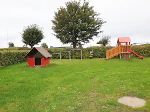 Spielplatz auf dem Campingplatz mit rotem Klettergerüst, Rutsche, kleinem Häuschen und Rasenfläche, Hecke und Bäumen im Hintergrund.
