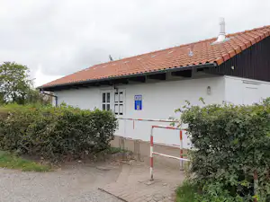 Campinghaus mit rotem Ziegeldach, weißer Fassade und Eingangstür, davor gepflasterter Weg und Hecken.
