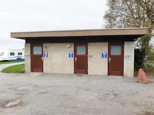Campinggebäude mit Ziegel- und Klinkerfassade, drei braunen Türen, Hinweisschildern und Kiesweg vor dem Eingang.