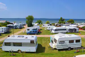 Reihen von Wohnwagen und Campingbussen auf einem gepflegten Stellplatz, im Hintergrund Wasser und Bäume am Ufer.
