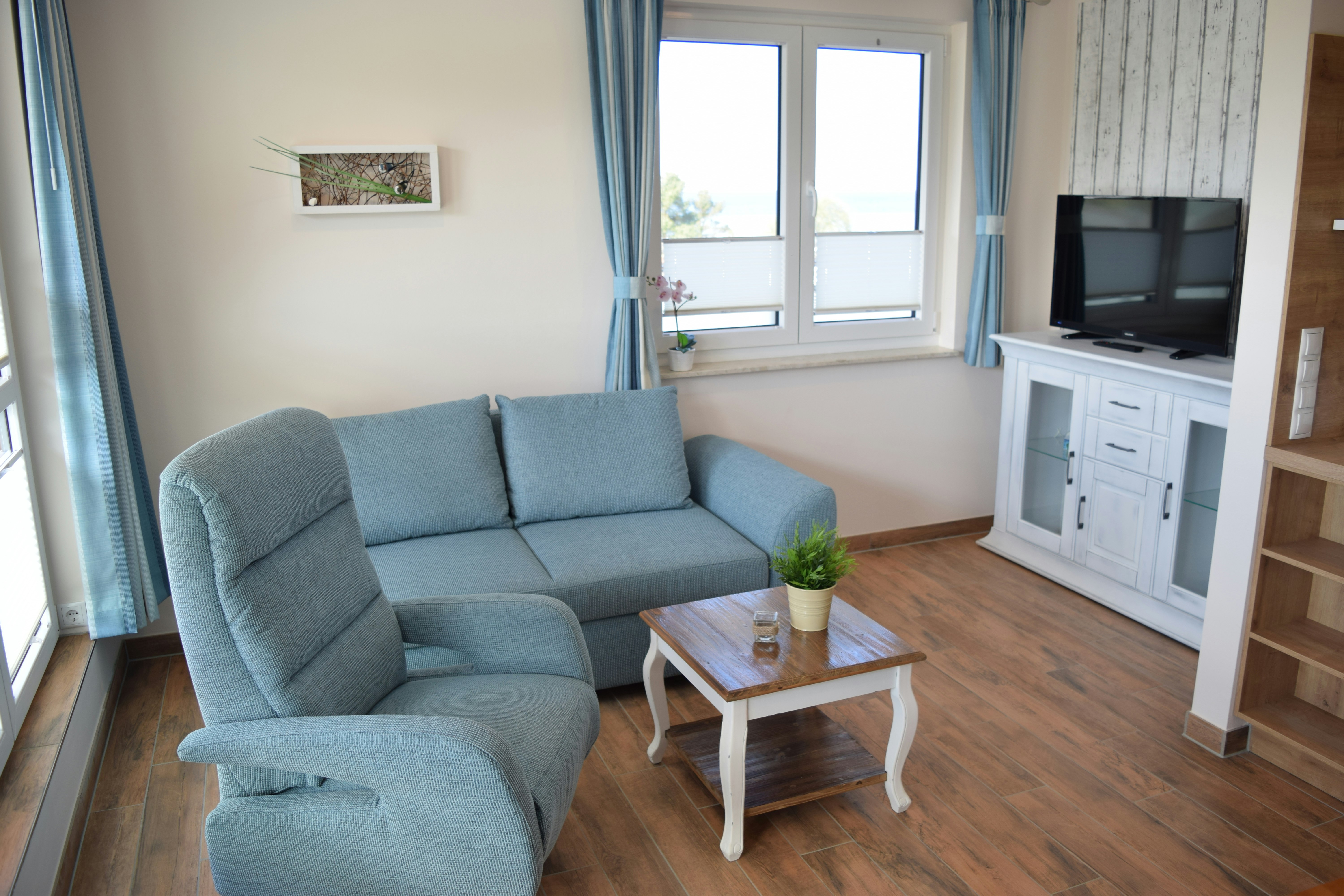 Blaues Sofa und weißer Couchtisch im hellen Wohnbereich mit Holzfußboden, Fensterfront und TV-Schrank.