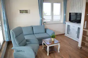Blaues Sofa und weißer Couchtisch im hellen Wohnbereich mit Holzfußboden, Fensterfront und TV-Schrank.