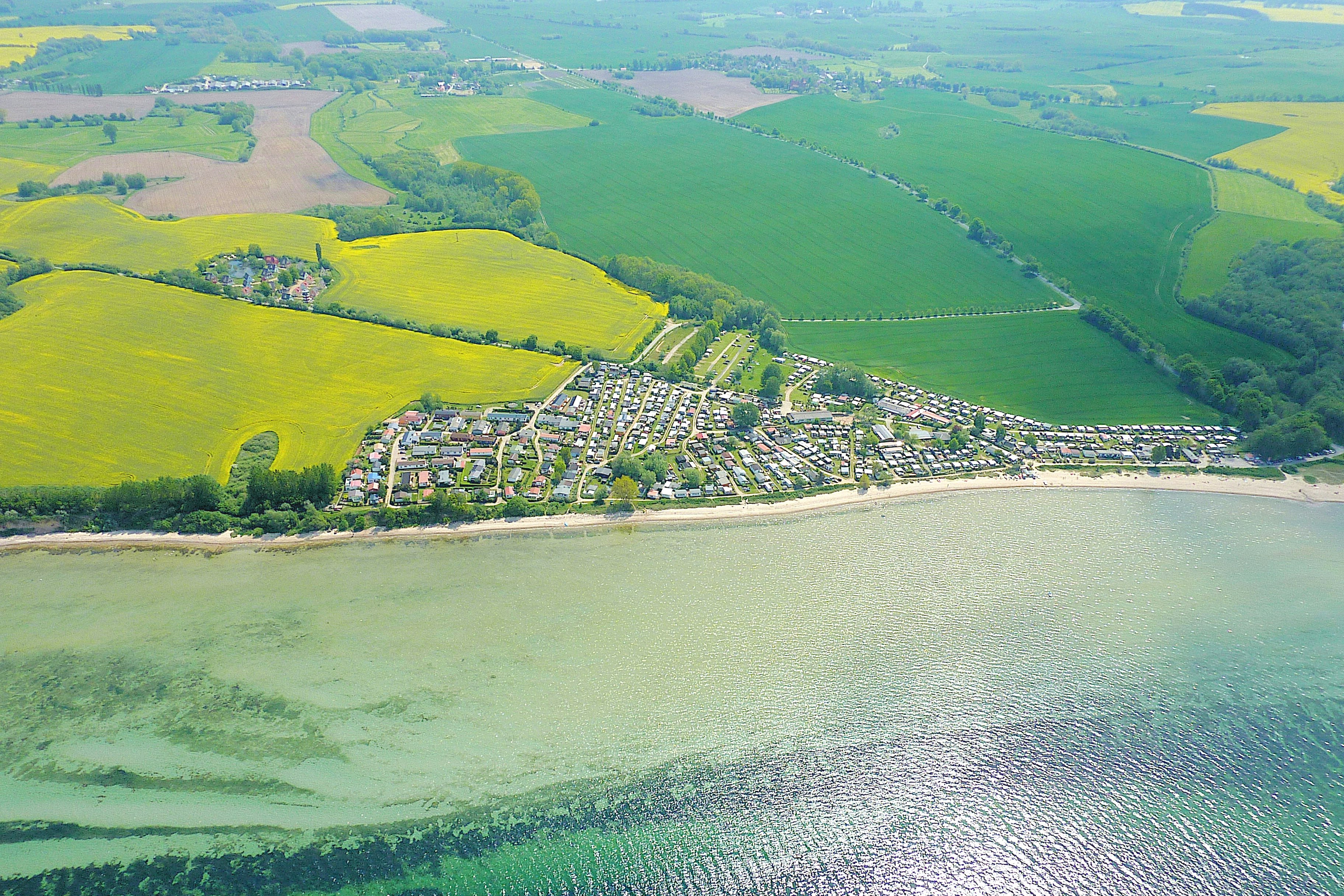Luftaufnahme eines Sees mit Uferlinie, dahinter ein Campingplatz mit vielen Stellplätzen und grünen Hügeln.