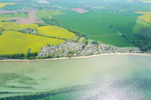 Luftaufnahme eines Sees mit Uferlinie, dahinter ein Campingplatz mit vielen Stellplätzen und grünen Hügeln.