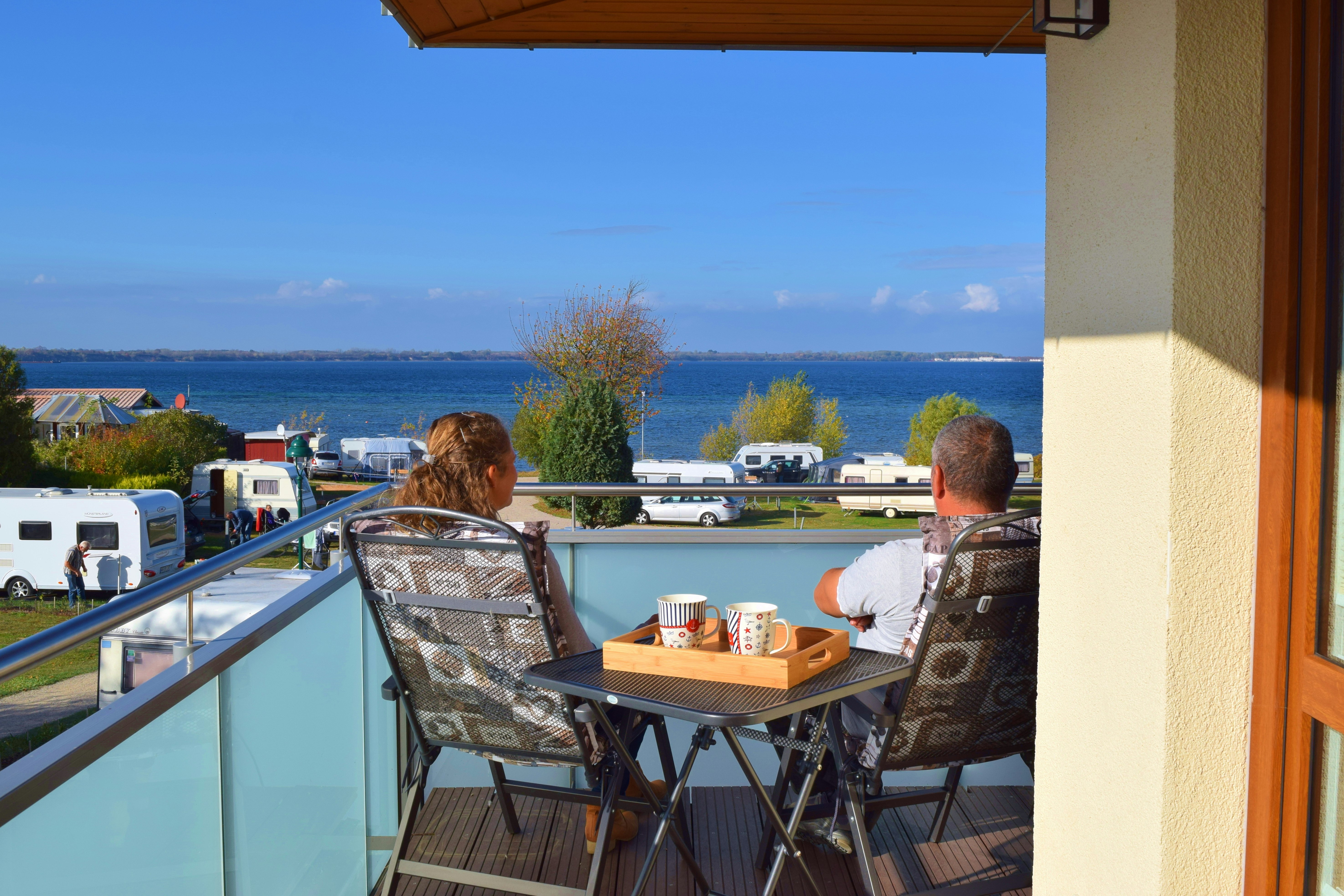 Balkon mit Tisch und Stühlen, Blick auf den See und Wohnwagen auf dem Campingplatz.