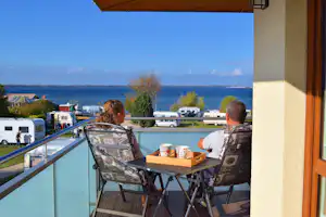 Balkon mit Tisch und Stühlen, Blick auf den See und Wohnwagen auf dem Campingplatz.
