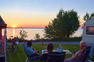 Camping am See bei Sonnenuntergang: Personen sitzen am Tisch vor dem Wohnwagen, Fahrräder und Bäume am Ufer.