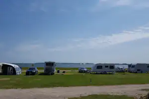 Campingplatz mit Wohnwagen und Autos auf grünem Rasen, im Hintergrund das Meer und ein flacher Horizont.