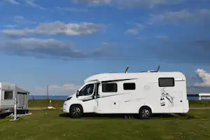 Weißes Wohnmobil auf einem Campingplatz am Meer, mit Rasenfläche, Zaun und einem weiteren Wohnwagen links.