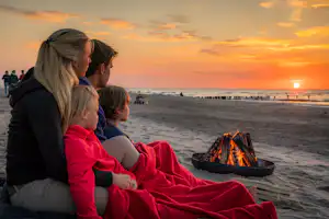 Familie sitzt am Strand bei Sonnenuntergang, Decke und Lagerfeuerkorb vor ihnen, Meer und Promenade im Hintergrund.