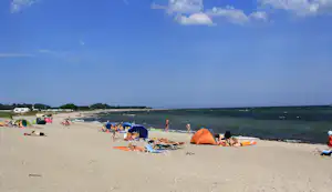 Strand mit Campingzelten, Liegestühlen und vielen Badegästen am Meer, im Hintergrund Wohnwagen und Küste.