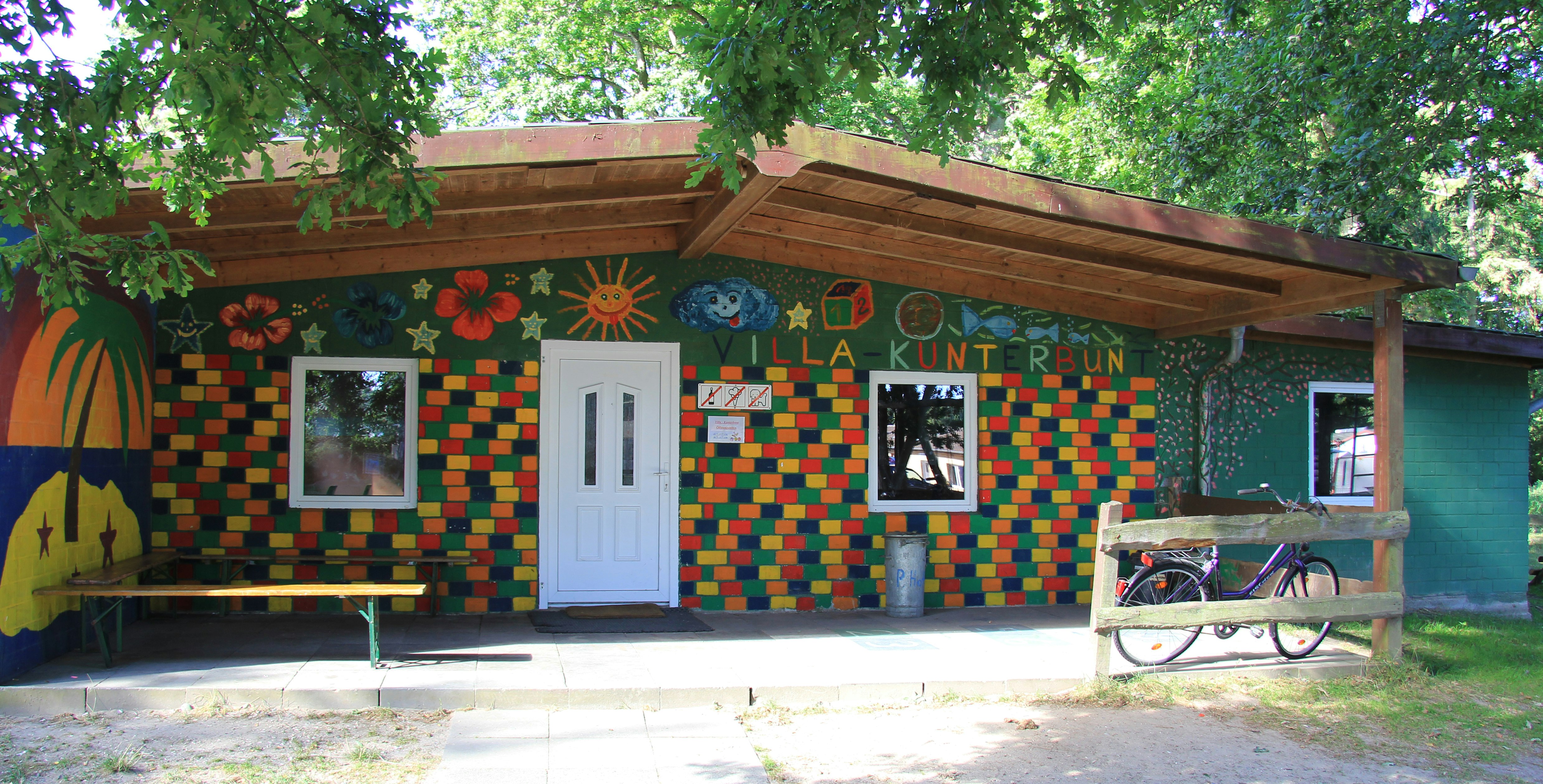 Bunt bemalte Campinghütte mit Holzvordach, zwei Fenstern und weißer Tür, davor Bank und Fahrrad im Schatten der Bäume.
