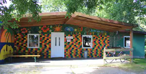 Bunt bemalte Campinghütte mit Holzvordach, zwei Fenstern und weißer Tür, davor Bank und Fahrrad im Schatten der Bäume.