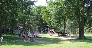 Spielplatz im Grünen mit Rutsche, Klettergerüst und Sandfläche zwischen hohen Bäumen auf dem Campinggelände.