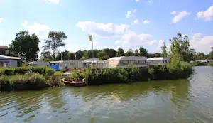 Wohnwagen und Caravans am Seeufer, mit Ruderboot im Wasser und Büschen im Vordergrund.