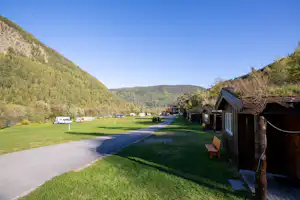 Campingplatz im Tal: asphaltierter Weg, Rasenflächen und kleine Hütten am Rand, im Hintergrund Berge und Bäume.