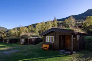 Holzhütte auf einem Campingplatz mit Rasenfläche, Bank und Holzschuppen im Hintergrund, umgeben von Bäumen und Hügeln.