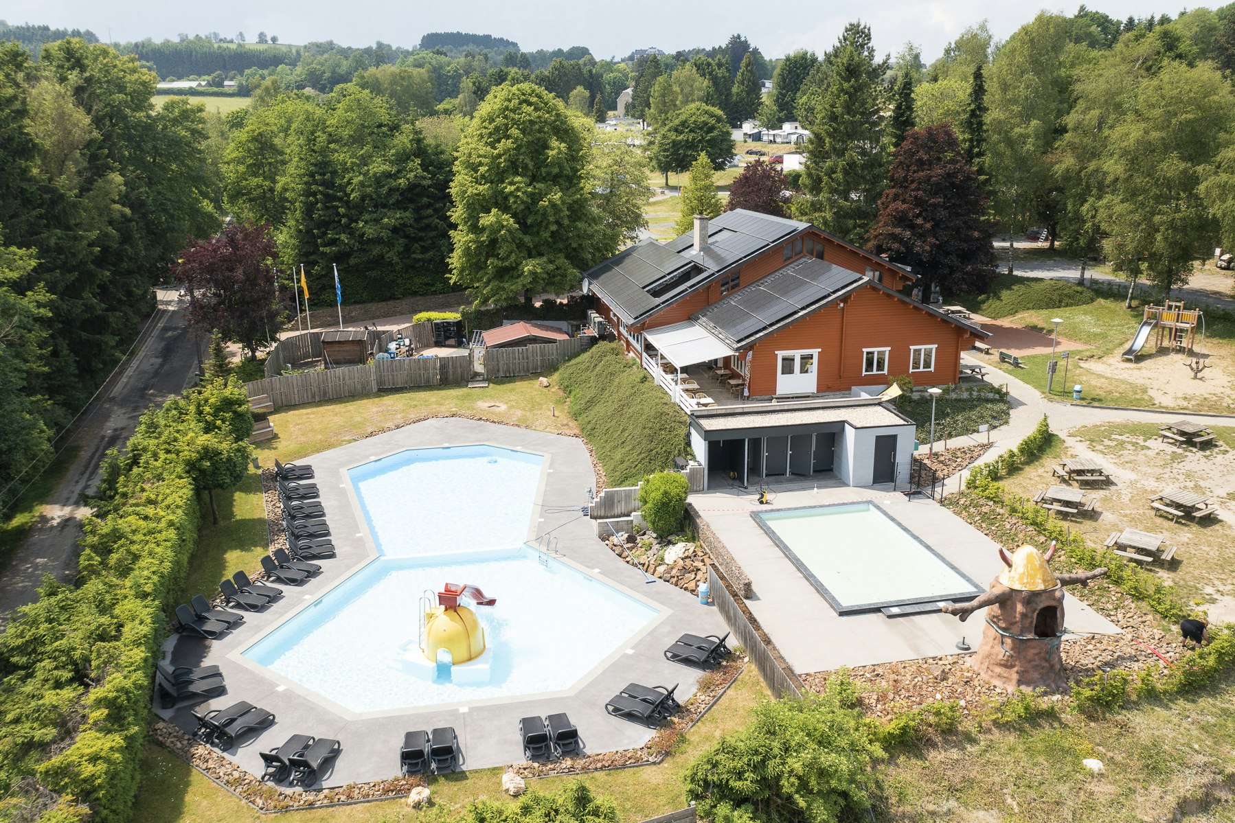 Ferienhaus mit Terrasse, zwei Sonnenliegen und großem Pool im Garten, umgeben von Bäumen und Wegen.