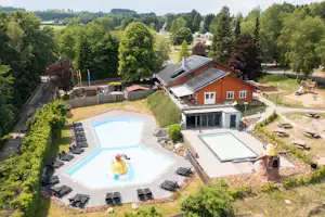 Ferienhaus mit Terrasse, zwei Sonnenliegen und großem Pool im Garten, umgeben von Bäumen und Wegen.