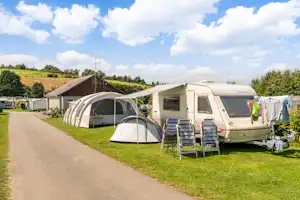 Wohnwagen und Vorzelt auf einem Campingplatz, daneben Stühle und Tisch auf dem Rasen, Weg entlang der Stellplätze.
