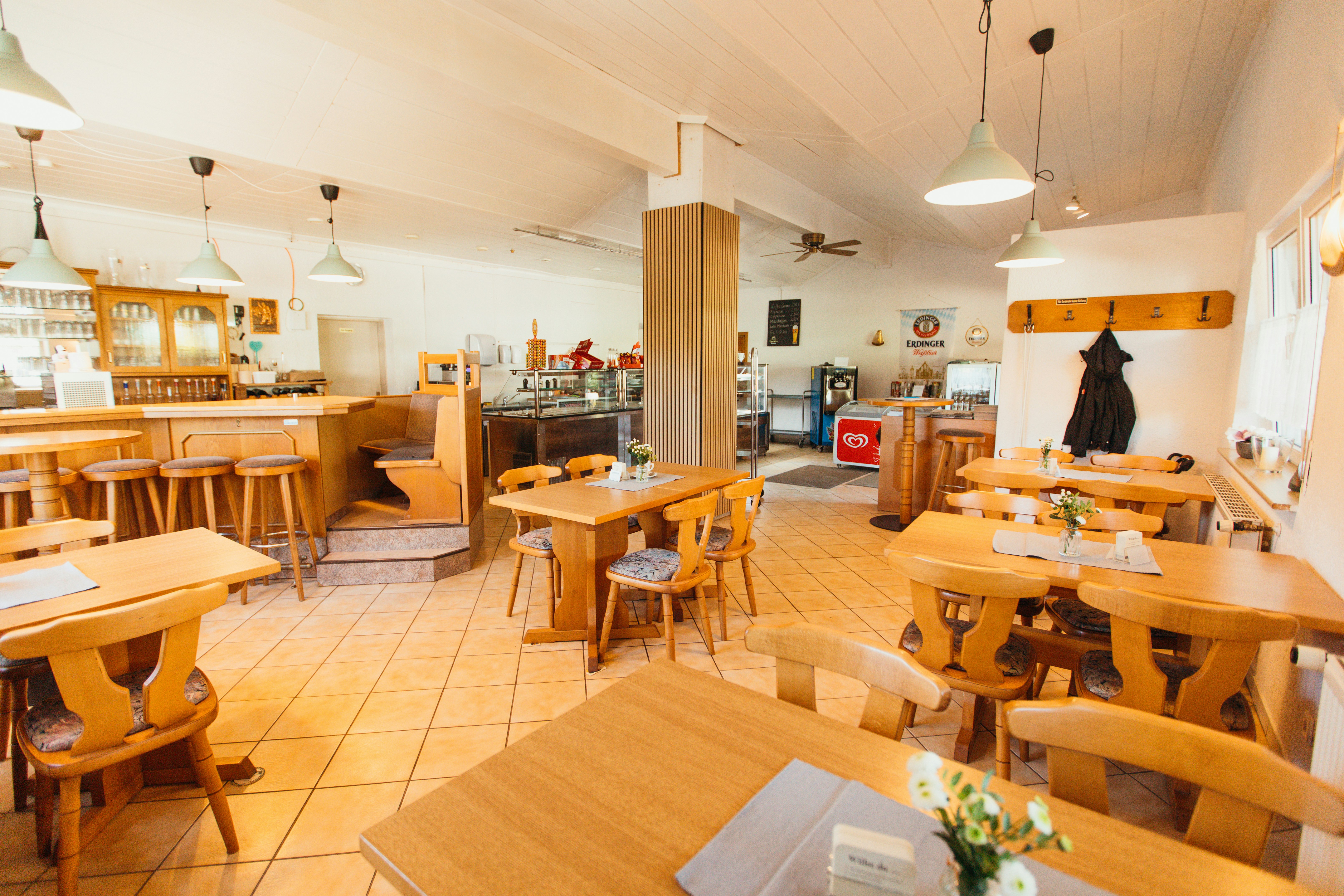 Innenraum einer gemütlichen Camping-Lounge mit Holztischen, Stühlen, Barbereich und großen Fenstern, helles Licht.