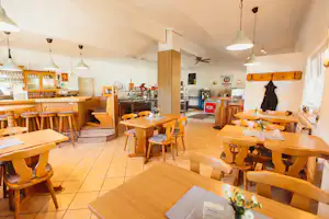 Innenraum einer gemütlichen Camping-Lounge mit Holztischen, Stühlen, Barbereich und großen Fenstern, helles Licht.