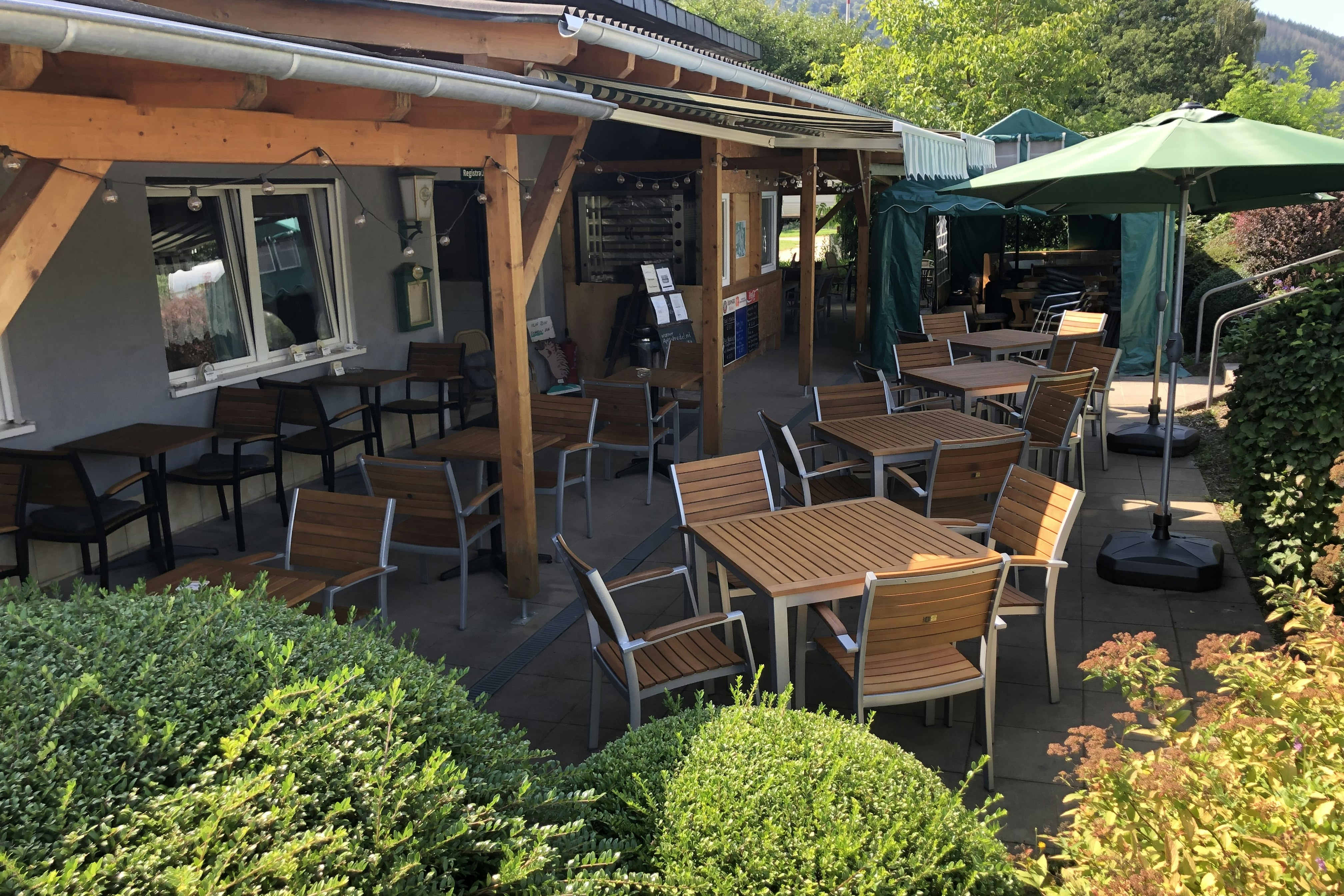 Terrasse am Campingplatz mit Holzdach, mehreren Gartenstühlen und Tischen sowie einem grünen Sonnenschirm, umgeben von Büschen und Hecken.