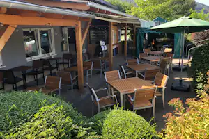 Terrasse am Campingplatz mit Holzdach, mehreren Gartenstühlen und Tischen sowie einem grünen Sonnenschirm, umgeben von Büschen und Hecken.