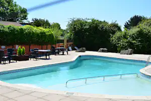 Schwimmbecken eines Campingplatzes mit Liegen und Tischen im Garten, Hecken und Bäume im Hintergrund.