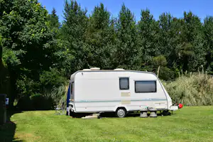 Weißer Wohnwagen auf einem grünen Stellplatz, umgeben von hohen Bäumen und dichtem Buschwerk im Campingbereich.