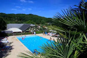 Blauer Camping-Pool mit Liegen und Sonnenschirmen, im Vordergrund Palmen, dahinter Gebäude und grüne Hügel.