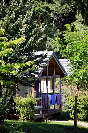 Holzunterkunft mit Veranda und Sitzbank im Garten, umgeben von Bäumen und Sträuchern.