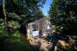 Wohnwagen mit Terrasse und Markise auf einem Campingplatz im Grünen, umgeben von Bäumen und einem gepflasterten Weg.