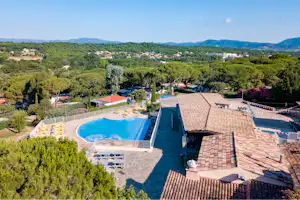 Campingpool mit Liegestühlen, gepflasterter Terrasse und Blick ins Grüne, im Hintergrund Häuser und Hügel.