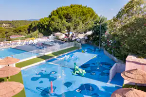 Großer Camping-Pool mit mehreren Becken, Sonnenschirmen und Liegen, umgeben von Bäumen und einem Gartenbereich.