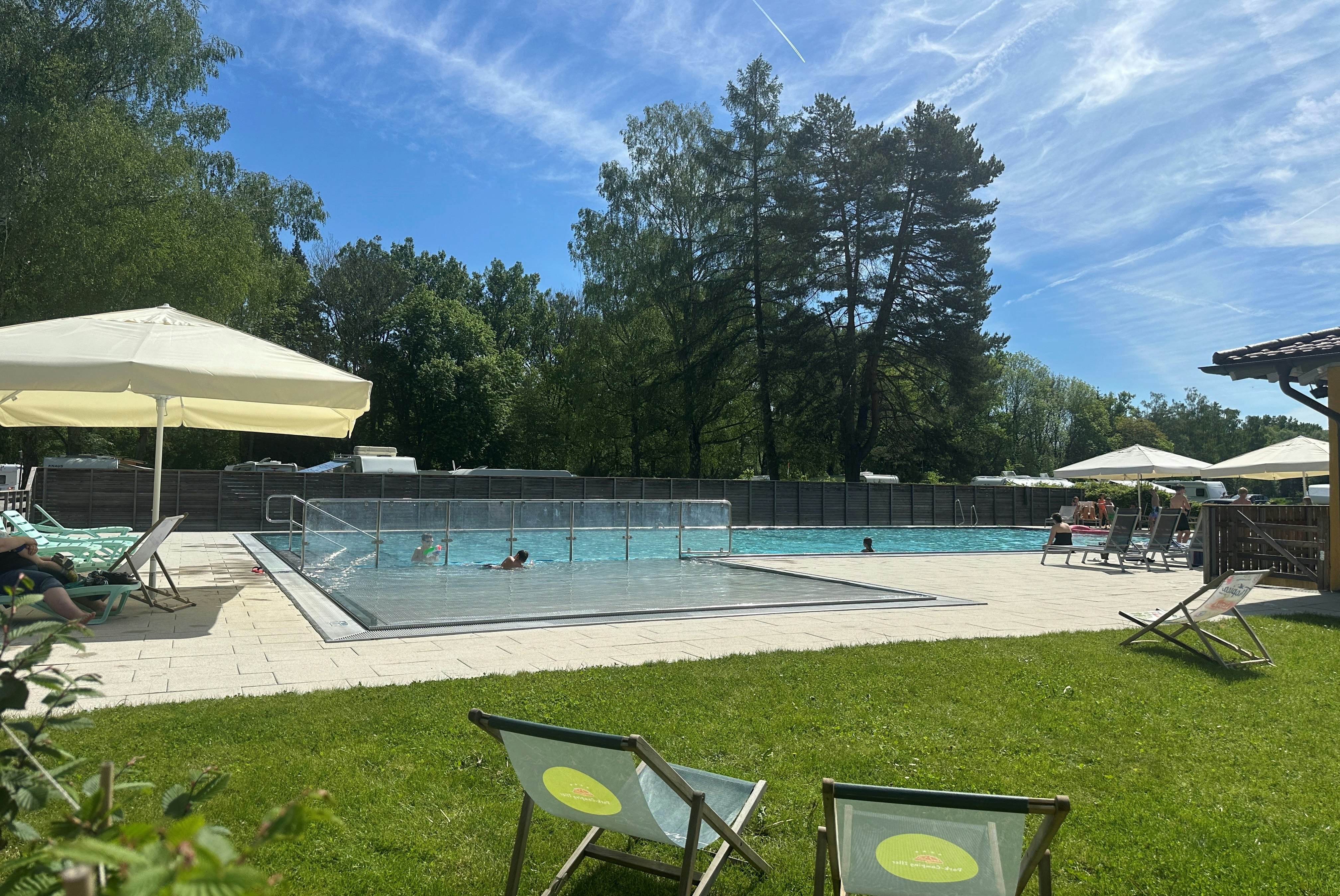 Schwimmbecken auf dem Campingplatz mit Sonnenschirmen, Liegestühlen und Blick auf Bäume und Wohnwagen im Hintergrund.