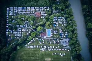 Luftaufnahme eines Campingplatzes mit vielen Stellplätzen, Wegen und Gebäuden am Fluss, umgeben von Bäumen.