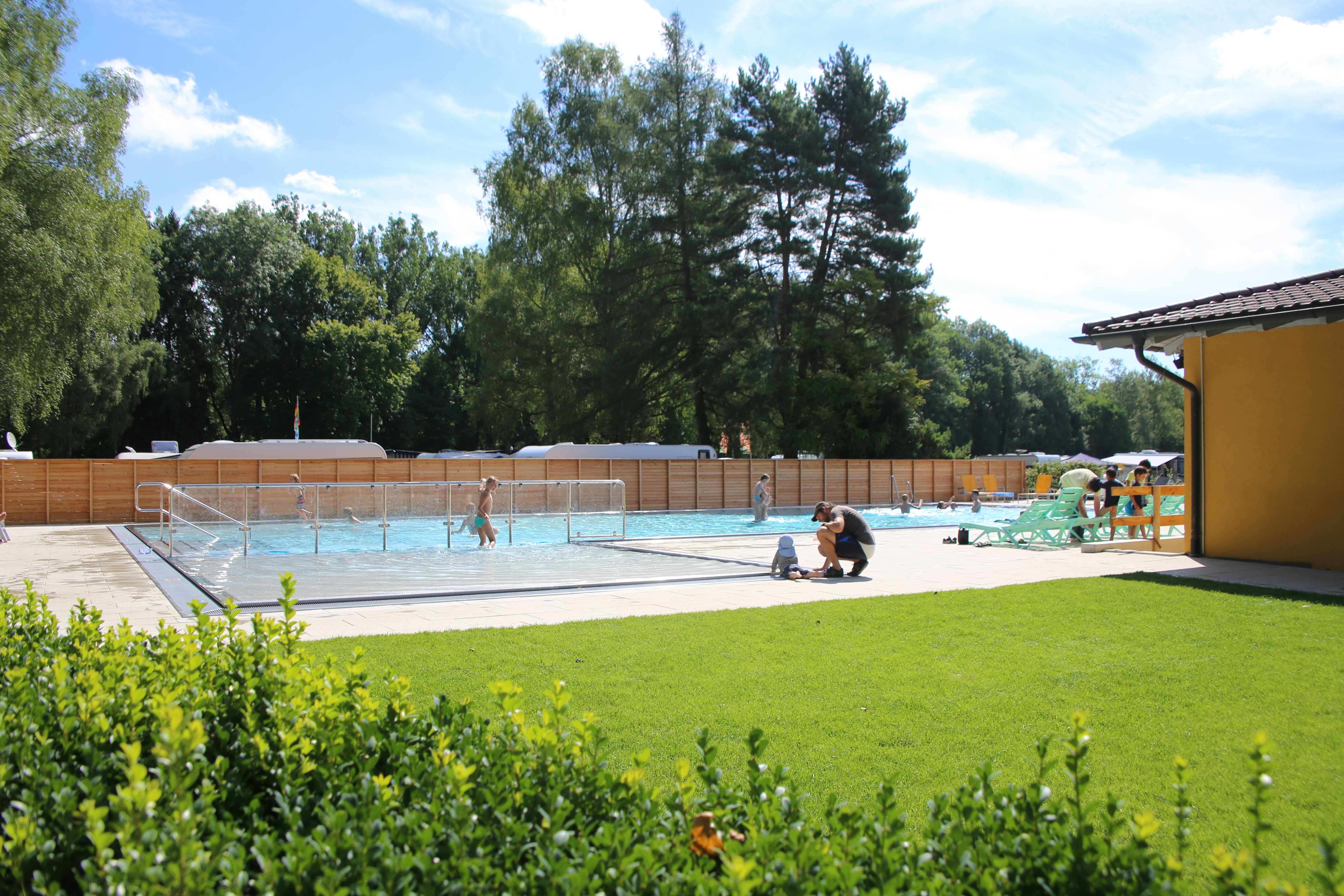 Schwimmbecken auf dem Campingplatz mit Treppe, Liegestühlen und Sonnenschirm am Rand, umgeben von Bäumen und Rasen.
