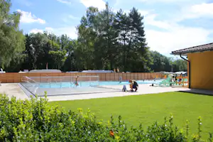 Schwimmbecken auf dem Campingplatz mit Treppe, Liegestühlen und Sonnenschirm am Rand, umgeben von Bäumen und Rasen.