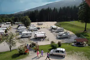 Wohnmobil-Stellplätze auf einem Kiesplatz, mehrere Camper und Autos, daneben Wiese und hohe Nadelbäume im Hintergrund.