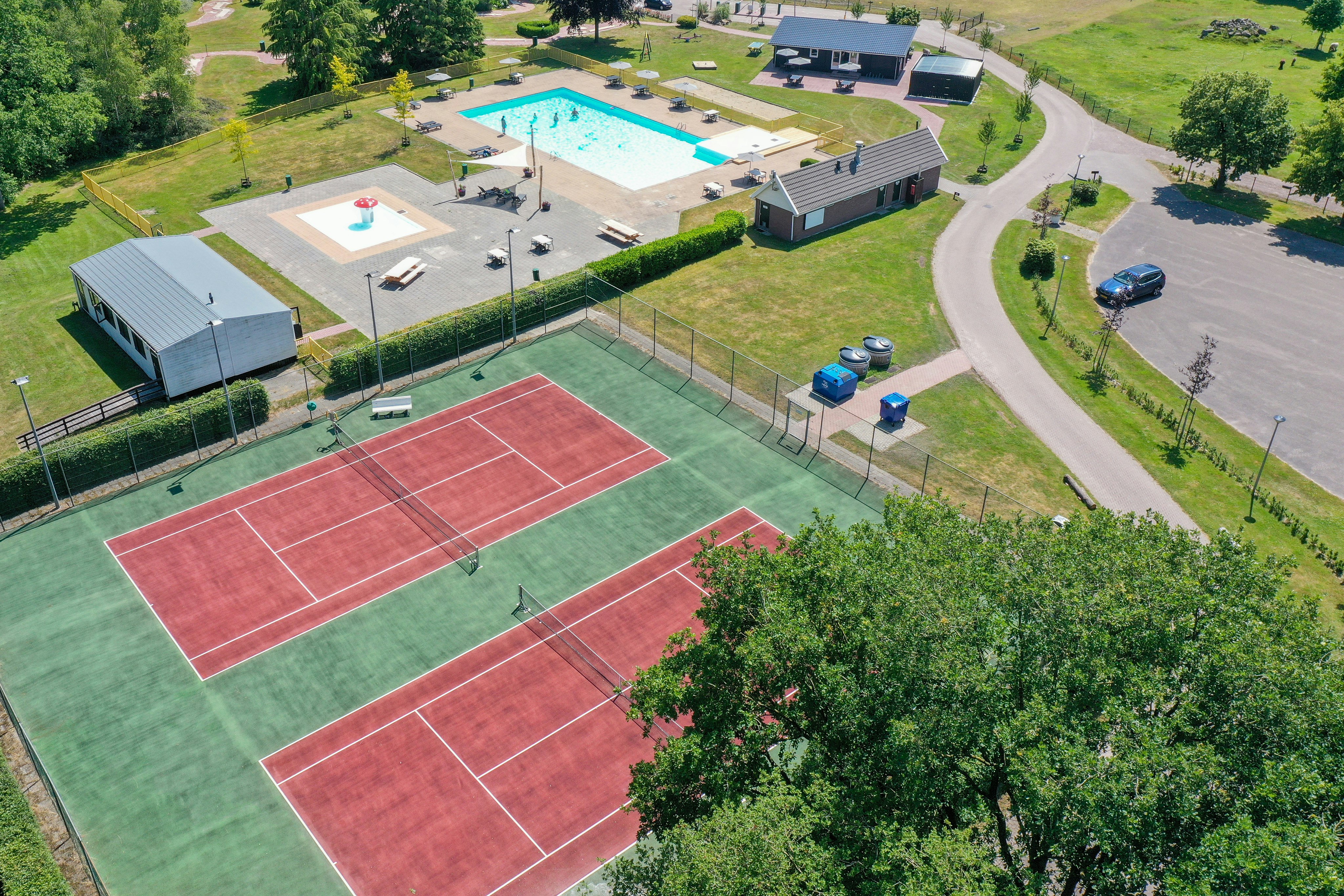Campinganlage mit großem Schwimmbecken, Tennisplätzen, mehreren Gebäuden, Terrasse und Wegen im grünen Gelände.