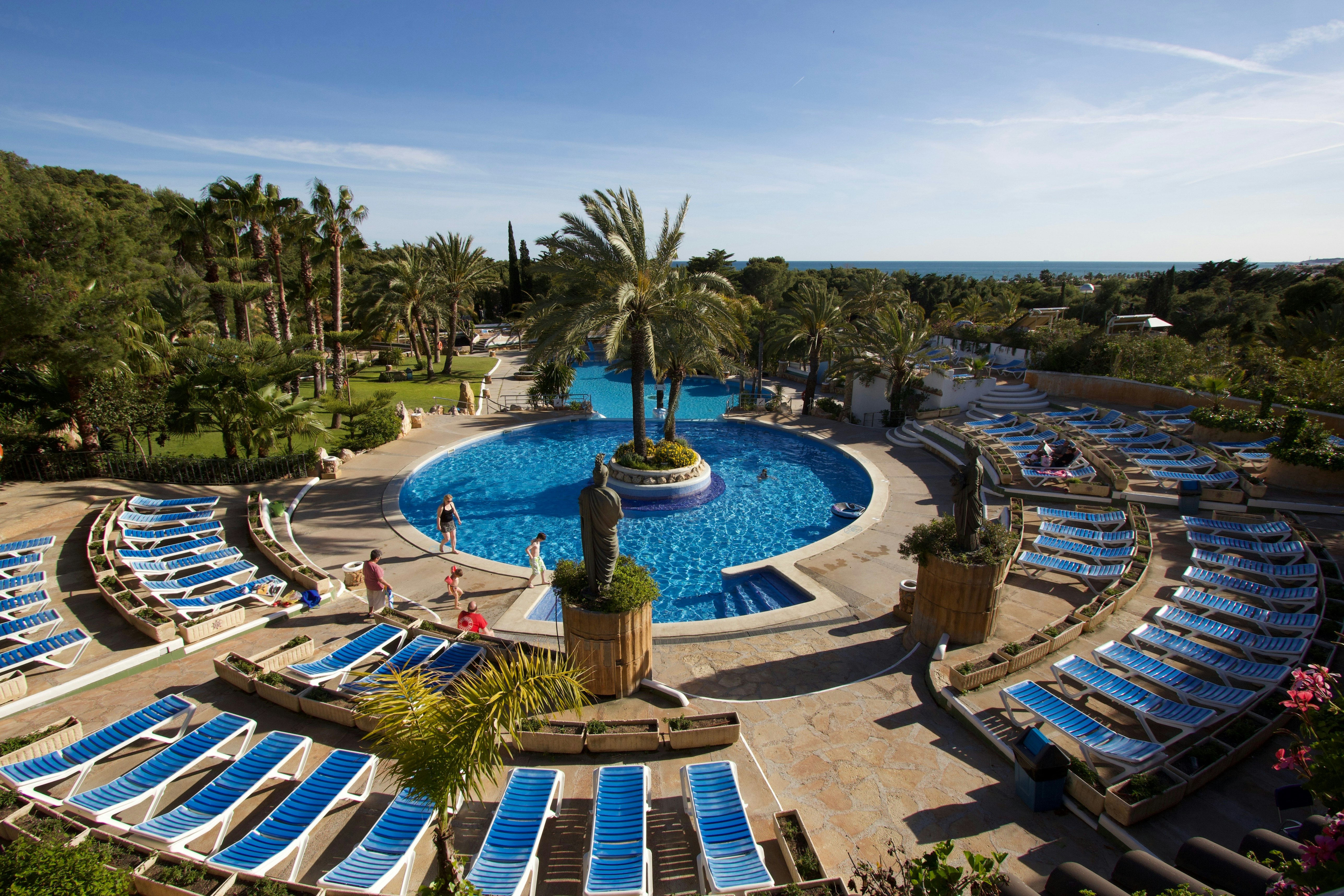 Camping-Pool mit rundem Becken, Palmen und vielen Sonnenliegen, Gartenweg und Blick auf das Meer im Hintergrund.