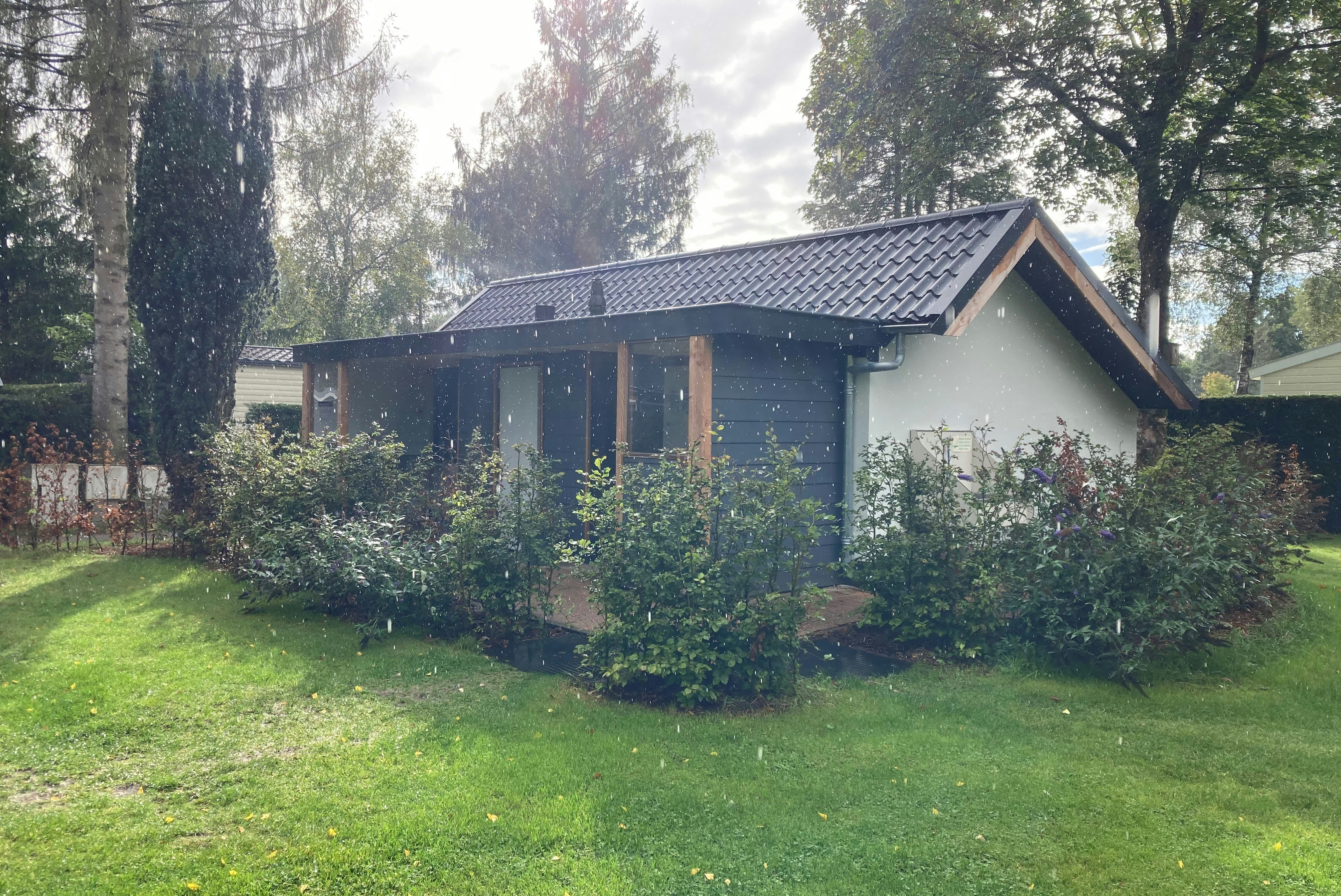 Campinghaus mit Satteldach und Veranda, umgeben von grünem Rasen, Büschen und hohen Bäumen im Garten.
