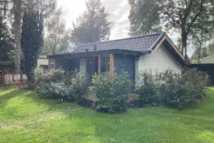 Campinghaus mit Satteldach und Veranda, umgeben von grünem Rasen, Büschen und hohen Bäumen im Garten.