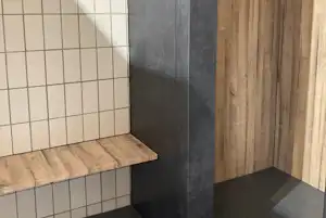 Duschbereich mit gefliesten Wänden, Holzverkleidung und grauem Boden, seitlich eine Armatur an der Wand.